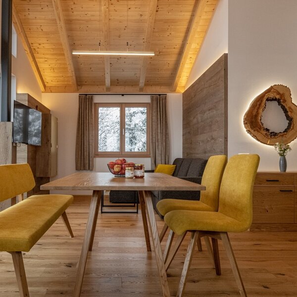 Der offene Ess- und Wohnbereich im Bauernhof mit einem Holztisch, gelben Sitzgelegenheiten, Sofa und großen Fenstern.