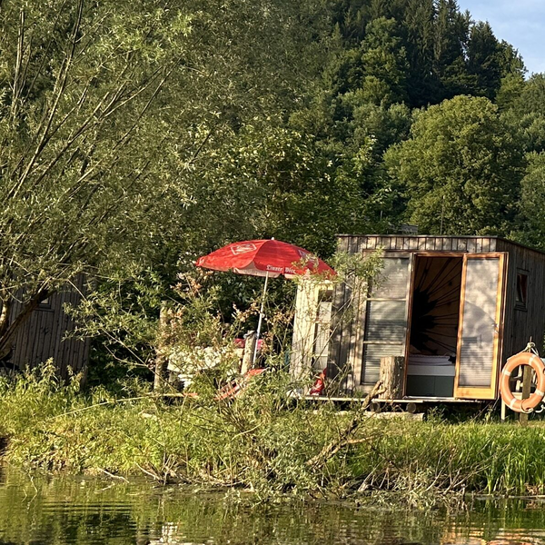 Schlafbox  mit Bett, rotem Sonnenschirm und Rettungsring am Wasser.