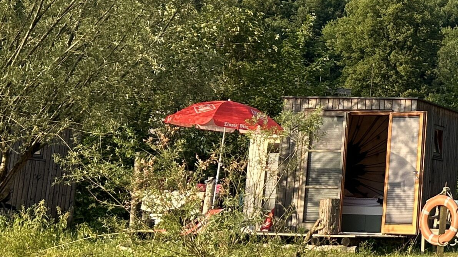 Schlafbox  mit Bett, rotem Sonnenschirm und Rettungsring am Wasser.