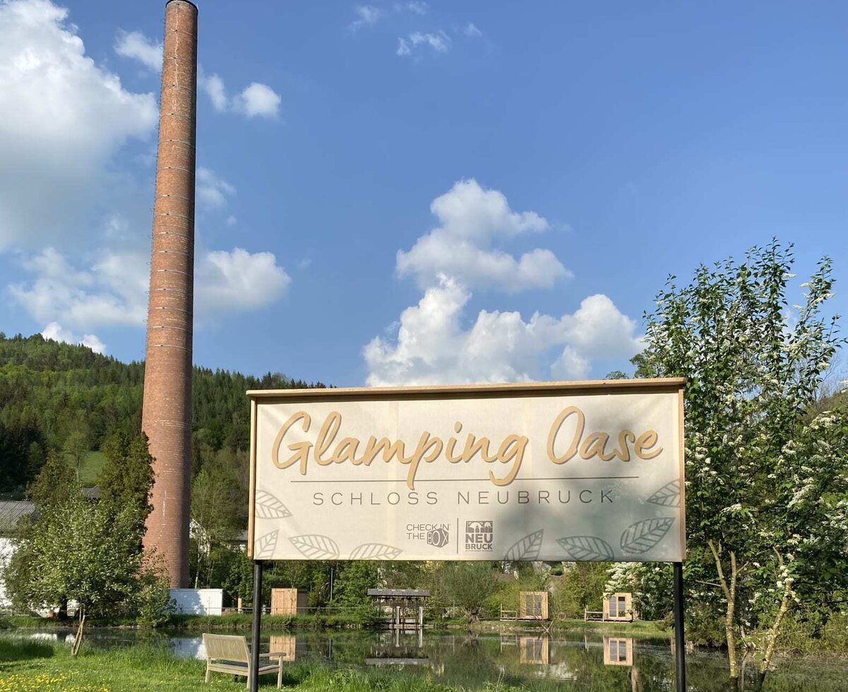 Die Glamping Oase  mit Schlafboxen rund um den Teich und einer hohen Esse (Schlot) im Hintergrund.