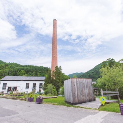 Das Gelände des Areals mit einem hohen Ziegelkamin (Schlot), altes Kesselhaus und grünen Hügeln im Hintergrund.