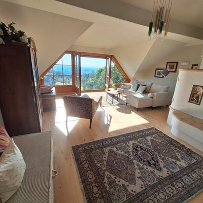 Das Wohnzimmer im Bauernhof ist ausgestattet mit einem Kachelofen, einem Sofa, einem Sessel, einem Holzschrank und großen Fenstern mit weitem Ausblick.