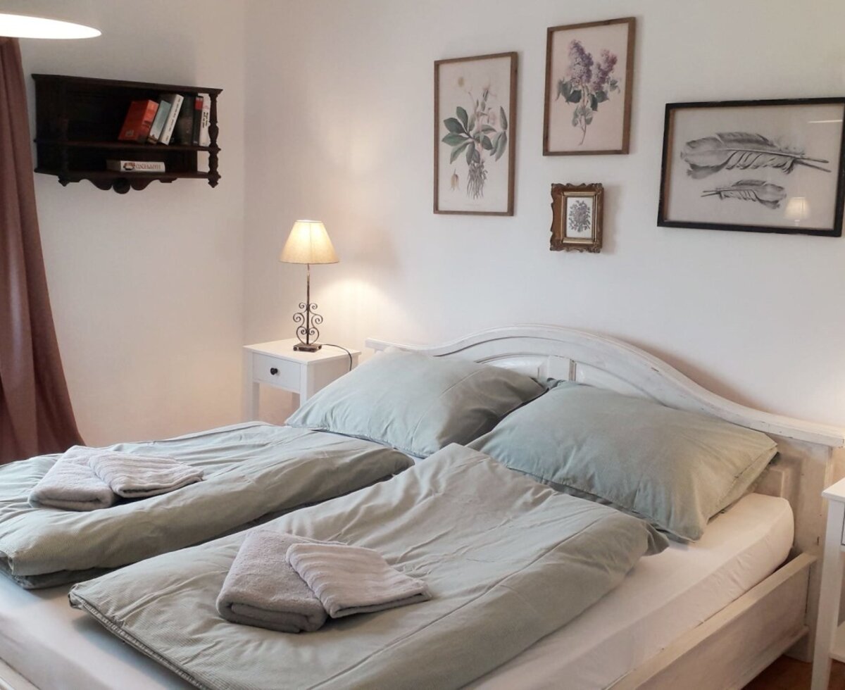 Schlafzimmer der Unterkunft mit Doppelbett, zwei Nachttischen, Leselampen, frischen Handtüchern und Fenster.