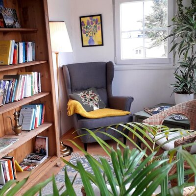 Leseecke in der Unterkunft mit Bücherregal, Sessel, Korbstuhl und Pflanzen.