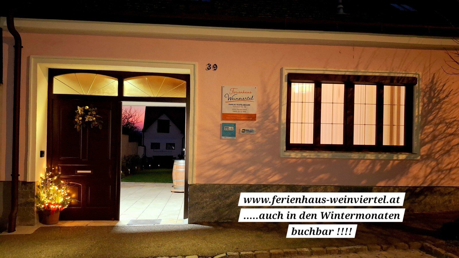 Die beleuchtete Fassade des Bauernhofs bei Nacht mit offenem Eingang, festlichen Dekorationen und Blick in den Garten.