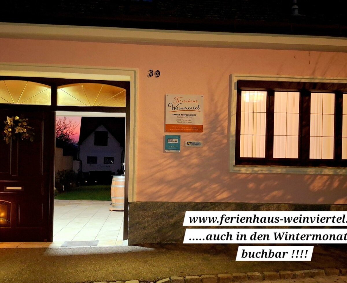 Die beleuchtete Fassade des Bauernhofs bei Nacht mit offenem Eingang, festlichen Dekorationen und Blick in den Garten.