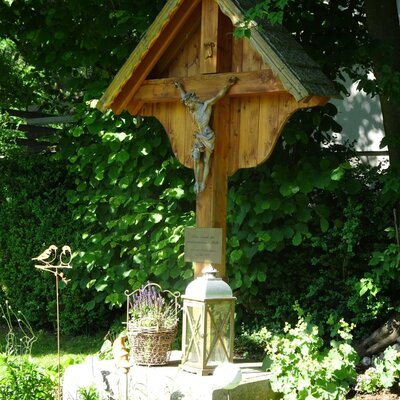 Das hölzerne Wegkreuz mit Kruzifix im Garten des Bauernhofs, dekoriert mit einem Lavendelkorb und einer Laterne.