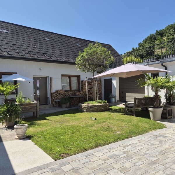 Der Innenhof des Bauernhofs mit einer Rasenfläche, gepflasterten Bereichen, Sonnenschirmen, einer Sitzbank und gestapeltem Brennholz am Gebäude.