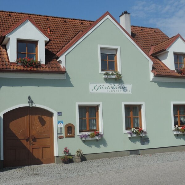 Die Außenansicht des Gästehauses am Bauernhof mit hellgrüner Fassade, roten Dachziegeln, großen Holztüren und blühenden Fensterkästen.
