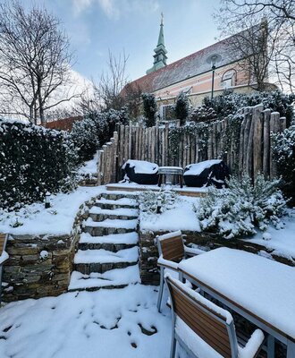 Der verschneite Gartenbereich des Weingenuss.Chalets mit einladenden Sitzgelegenheiten und Blick auf eine malerische Kirche im Hintergrund.