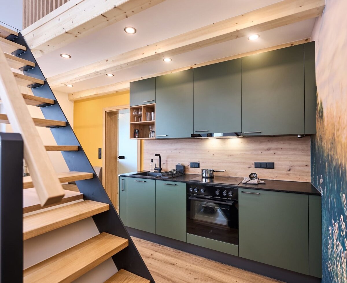Küche im Haus mit grünen Schränken, schwarzer Arbeitsplatte, Backofen und Herd, sowie einer Holztreppe.
