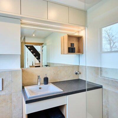 Badezimmer im Haus mit Waschbecken, beleuchtetem Spiegel, Wandschränken und einem Fenster.