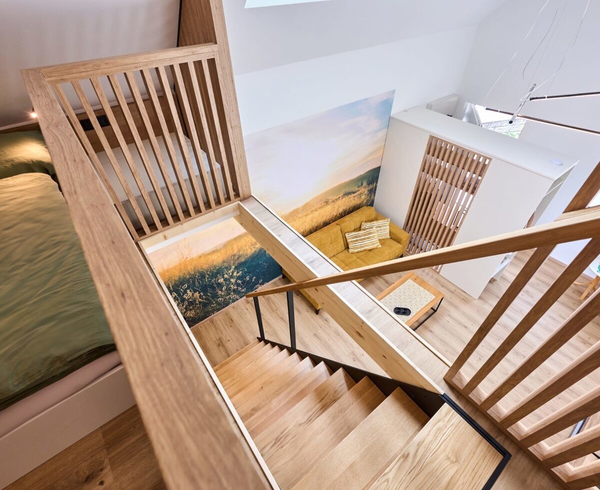 Innenansicht des Hauses mit Holztreppe, einem Schlafbereich auf der oberen Ebene und einem Wohnbereich mit gelbem Sofa und Wandgemälde auf der unteren Ebene.