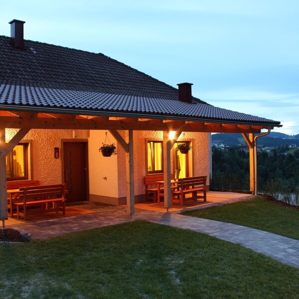 Die Außenansicht des Bauernhofs bei Dämmerung mit beleuchteter, überdachter Terrasse und Holzsitzgelegenheiten.
