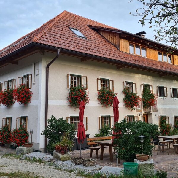 Der Bauernhof mit blühenden Geranien an den Fenstern und einem Außenbereich mit Holztischen und roten Sonnenschirmen.